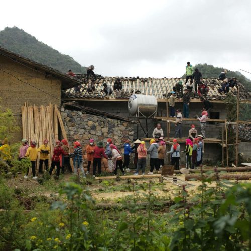 A group of people stands in front of a house under construction, with several individuals working on the roof. Large water tanks are visible next to the building, and hills are in the background.