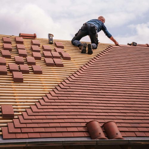 A person wearing work clothes installs tiles on a sloped roof. Stacks of tiles and tools are visible beside them, and part of the roof remains unfinished against a cloudy sky.