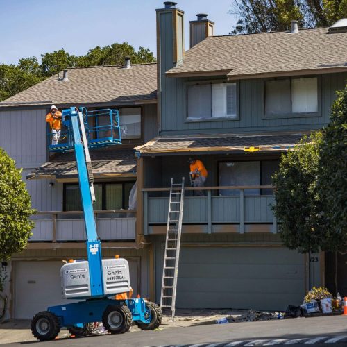 Two construction workers in orange shirts repair the roof of a gray two-story house using a blue hydraulic lift and a ladder. Tools and materials are scattered in the driveway below.