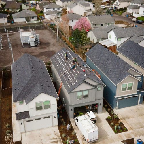 Aerial view of a residential neighborhood with workers installing solar panels on the roof of a two-story house. A white van is parked in the driveway, and equipment is visible on the ground and roof.
