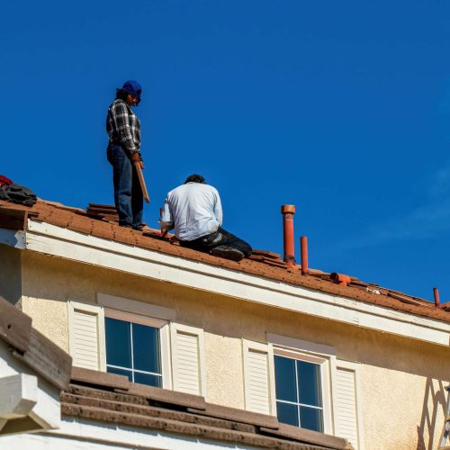 Two workers are repairing or installing shingles on the roof of a light-colored house under a clear blue sky. A ladder is leaning against the roof, and various tools are visible.