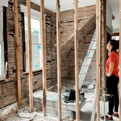 A woman in a red shirt and black pants stands in a partially demolished room with exposed wall studs, loose debris on the floor, and a silver ladder near the back wall. Sunlight comes through two uncovered windows.