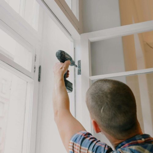 A person in a plaid shirt uses a cordless drill to fix or install the hinge of a white window frame in a well-lit room.