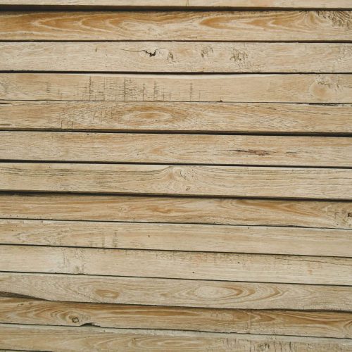 Close-up view of several horizontal wooden planks with visible grain patterns, knots, and natural imperfections. The wood is light brown and appears weathered.