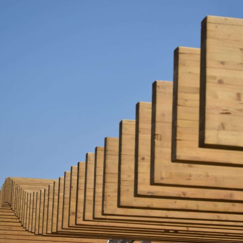 Series of rectangular wooden beams arranged in a repeating geometric pattern against a clear blue sky, creating a visually striking architectural structure.