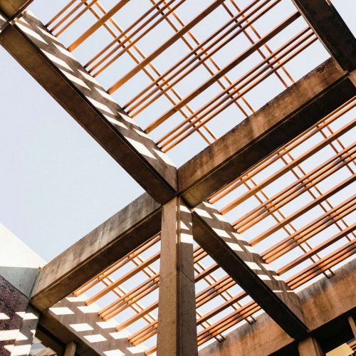 Open concrete structure with intersecting beams and a grid pattern overhead, casting geometric shadows on the columns below. The sky is visible through the gaps in the grid.