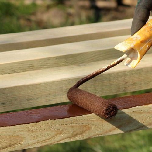 A gloved hand uses a paint roller to apply brown paint to a wooden plank, with unpainted planks visible in the background.