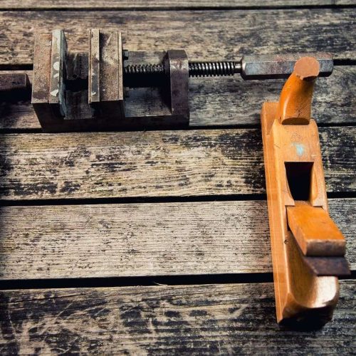 A hammer, a metal clamp, and a wooden hand plane are placed on a weathered wooden surface, arranged in a rough "L" shape.