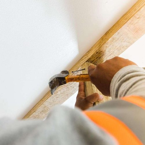 A person uses a hammer to drive a nail into a wooden board attached to a white wall. The person is wearing a gray shirt and an orange safety vest.