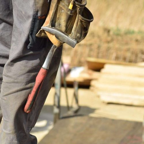 A person wearing gray work pants and a tool belt is standing outdoors, holding a hammer with a red handle. Construction materials are visible in the blurred background.